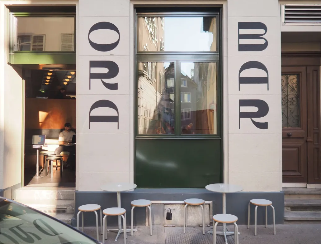ORA Bar storefront with ORA BAR lettering, green trim and pavement stools