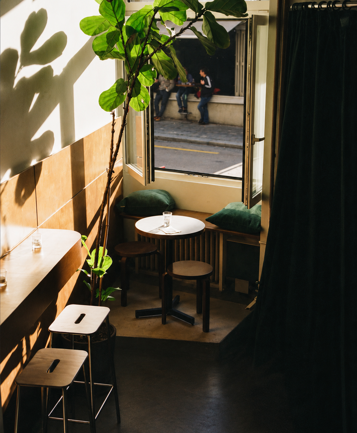 A sunlit window nook at ORA Bar with green velvet cushions, a fiddle-leaf plant and timber stools